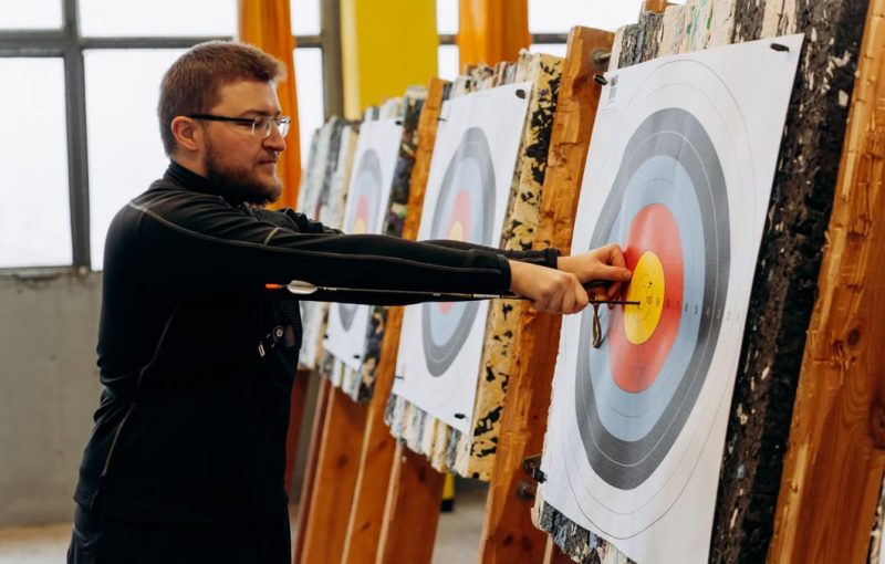 Man in glasses practicing archery indoors, pulling arrows from bullseye on target.